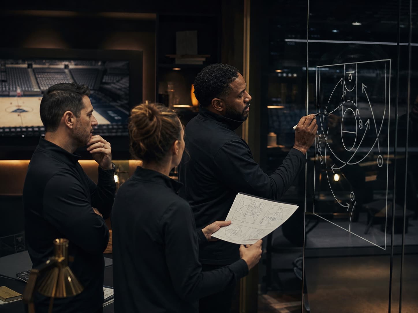 Basketball strategy staff drawing a half-court plan across a glass tactics wall